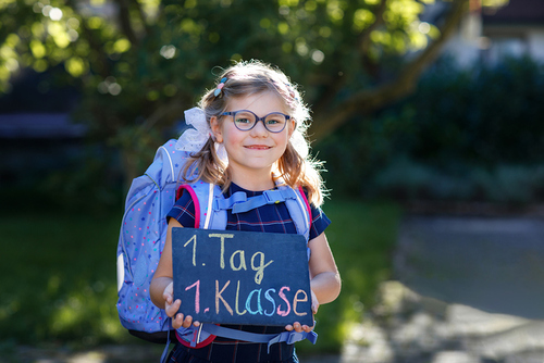 Happy little kid girl with eye glasses with backpack or satchel and big school bag on the first day of school. Healthy adorable child outdoors. Child holding chalks desk for first schoolday. / IRINA SCHMIDT, romrodinka Happy little kid girl with eye glasses with backpack or satchel and big school bag on the first day of school. Healthy adorable child outdoors. Child holding chalks desk for first schoolday